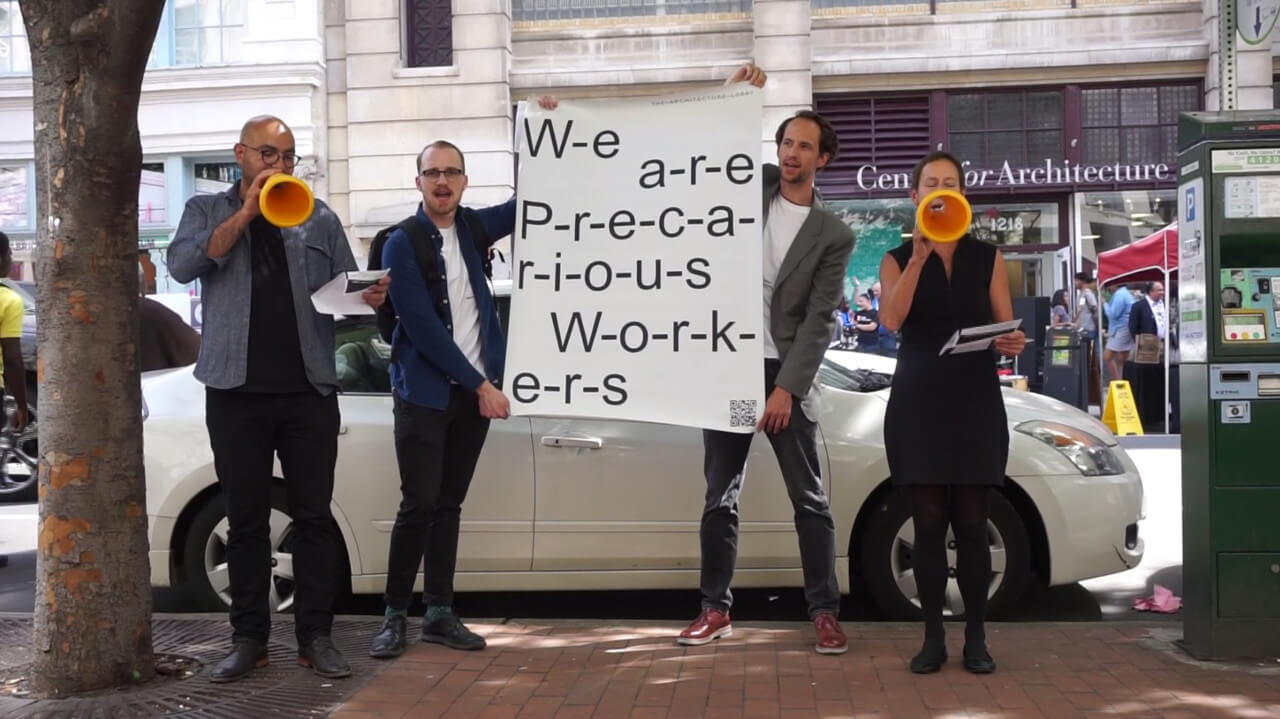 A group of architects outside of a building holding a "we are precarious workers" sign, ostensibly protesting the Trump administration