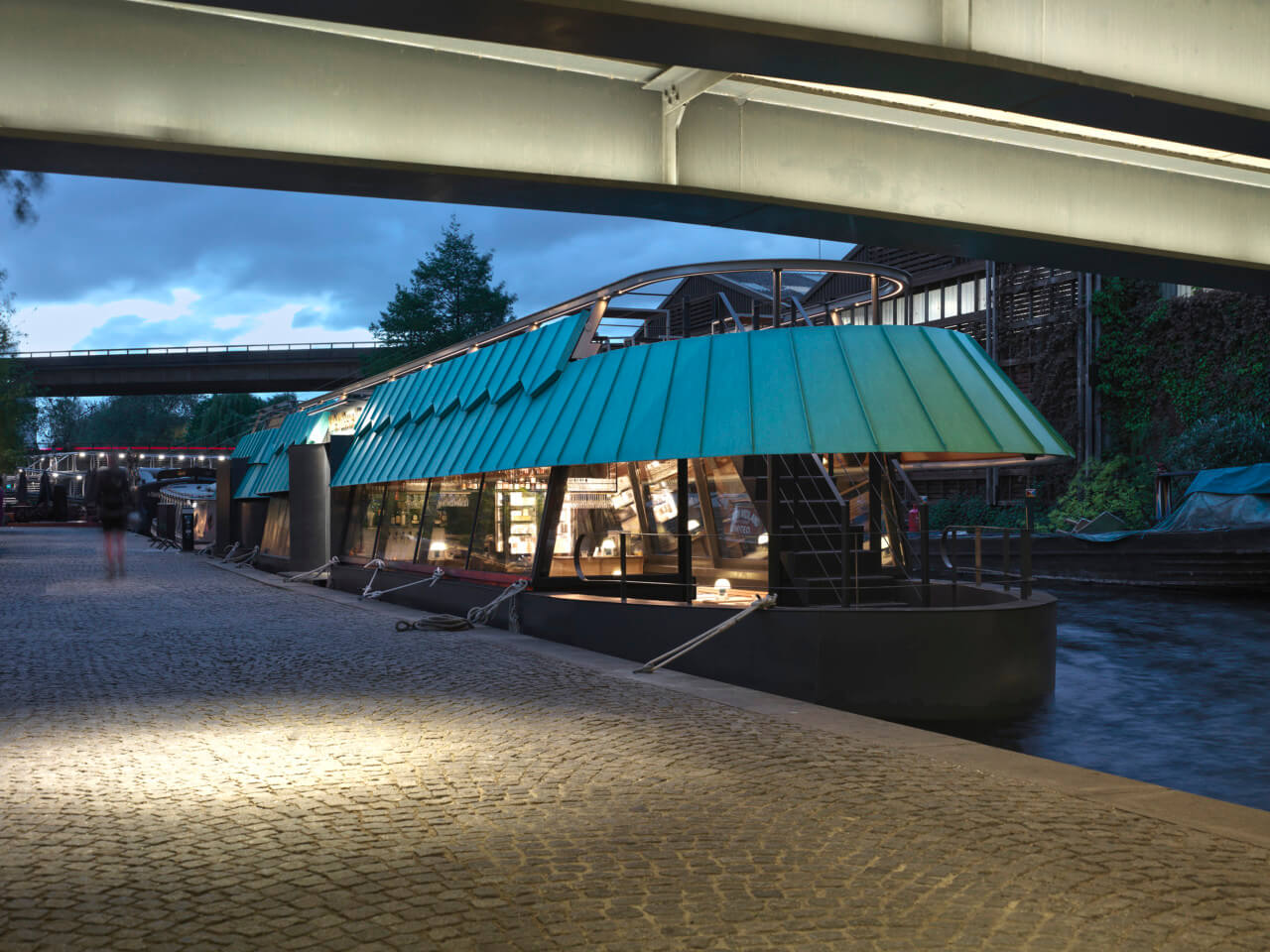 a barge-restaurant with a patinated roof as seen beneath a bridge at night