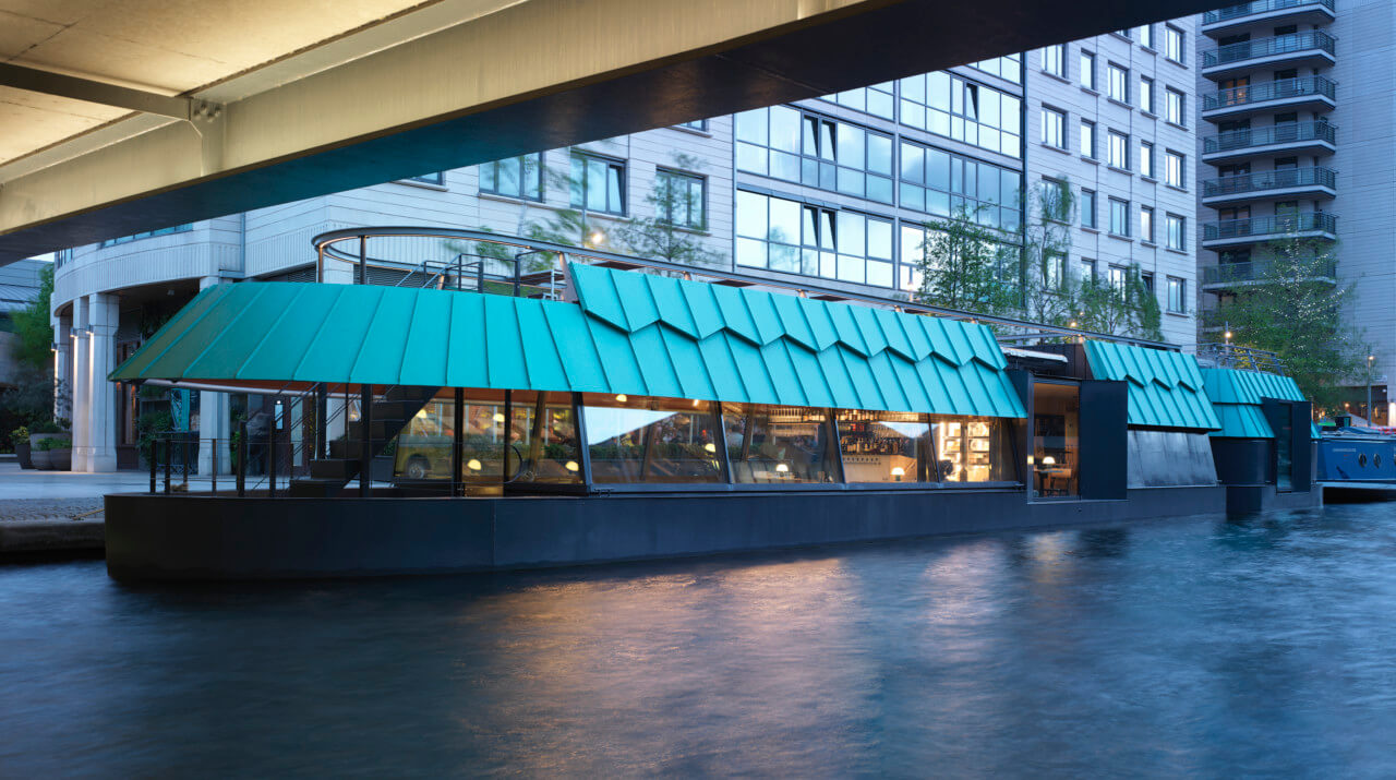 a barge-restaurant with a patinated roof as seen beneath a bridge