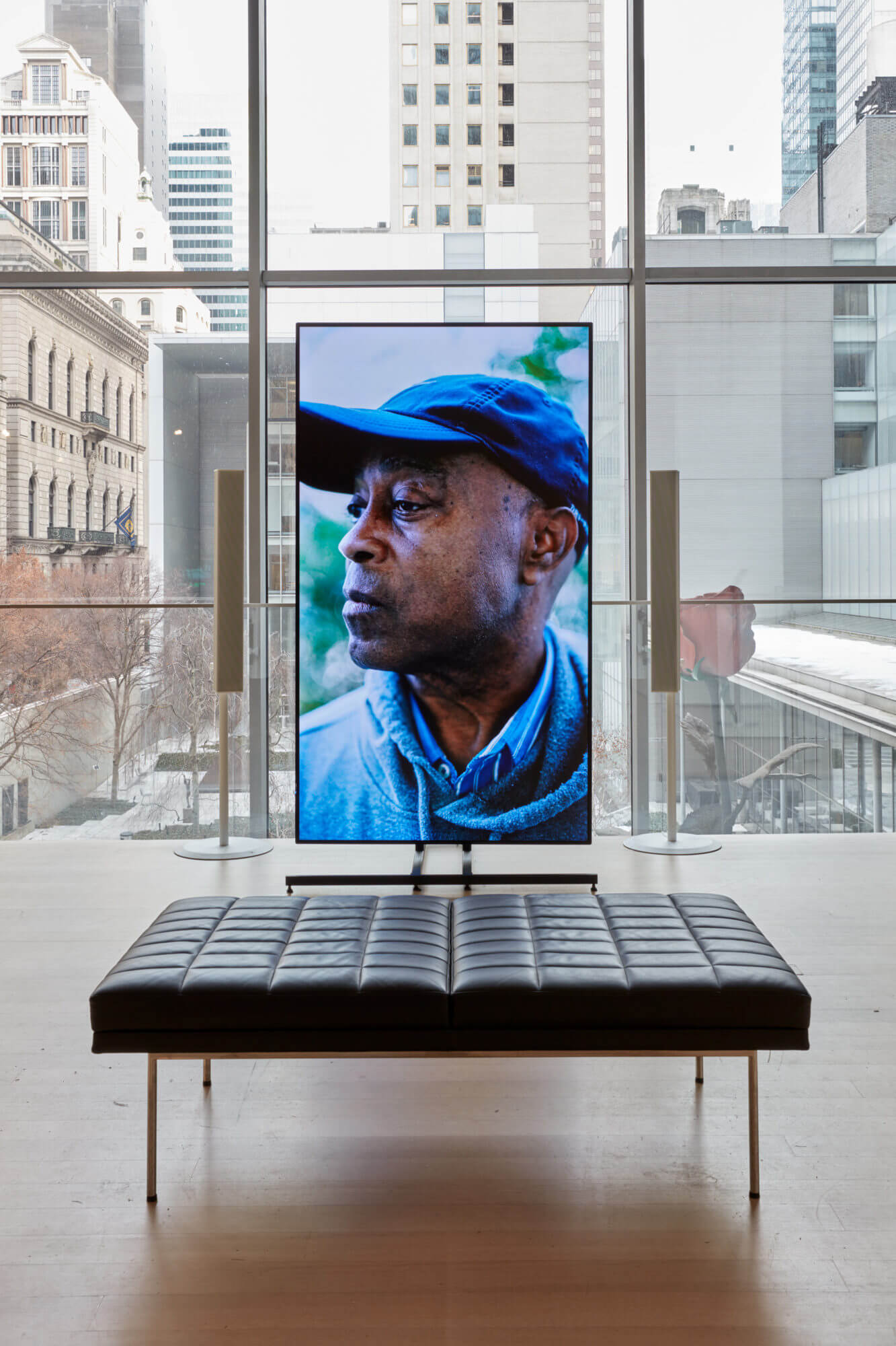photograph depicting a vertically oriented video screen set within a museum gallery with a cityscape in the background
