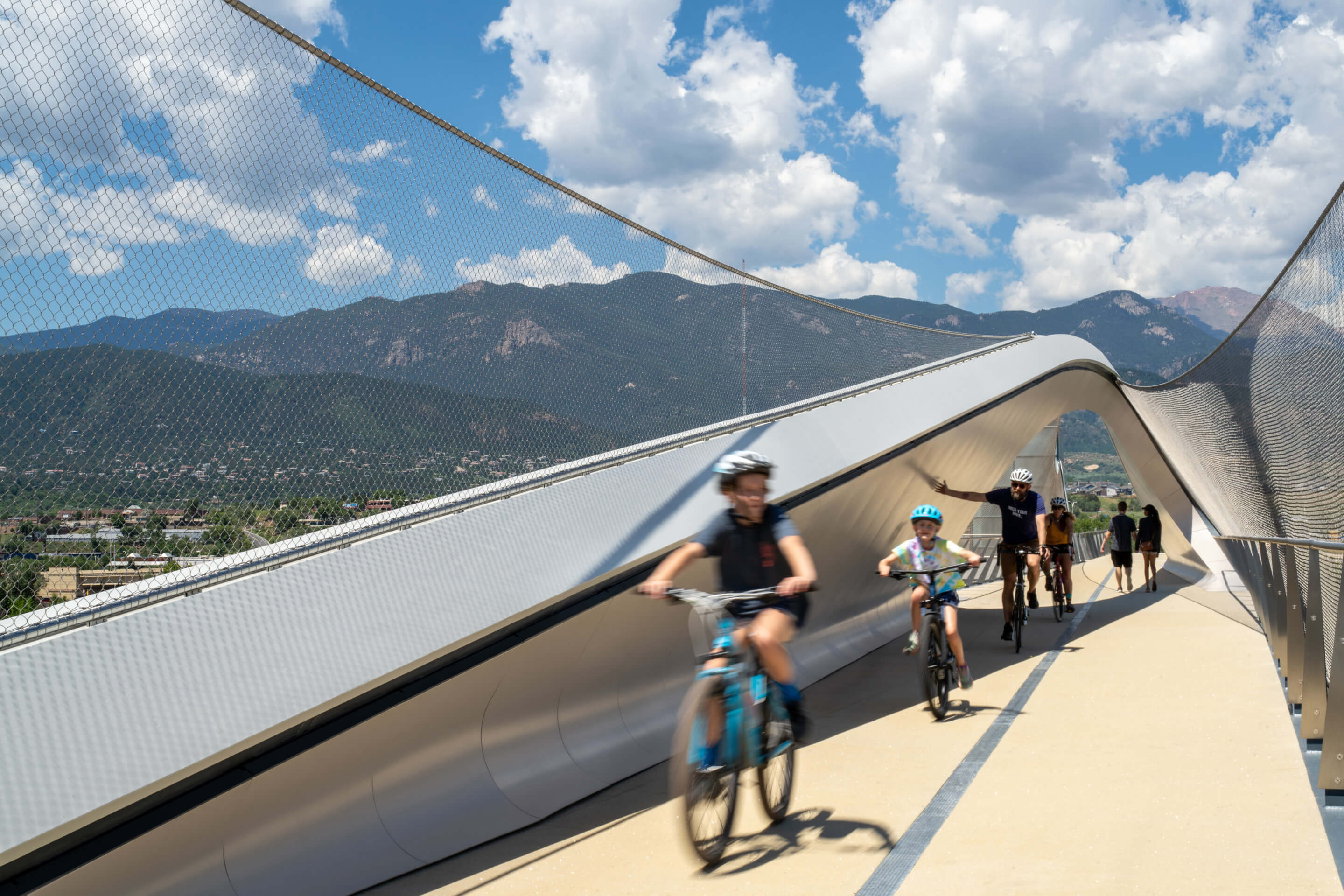 cyclists cross a pedestrian bridge with mountains in background