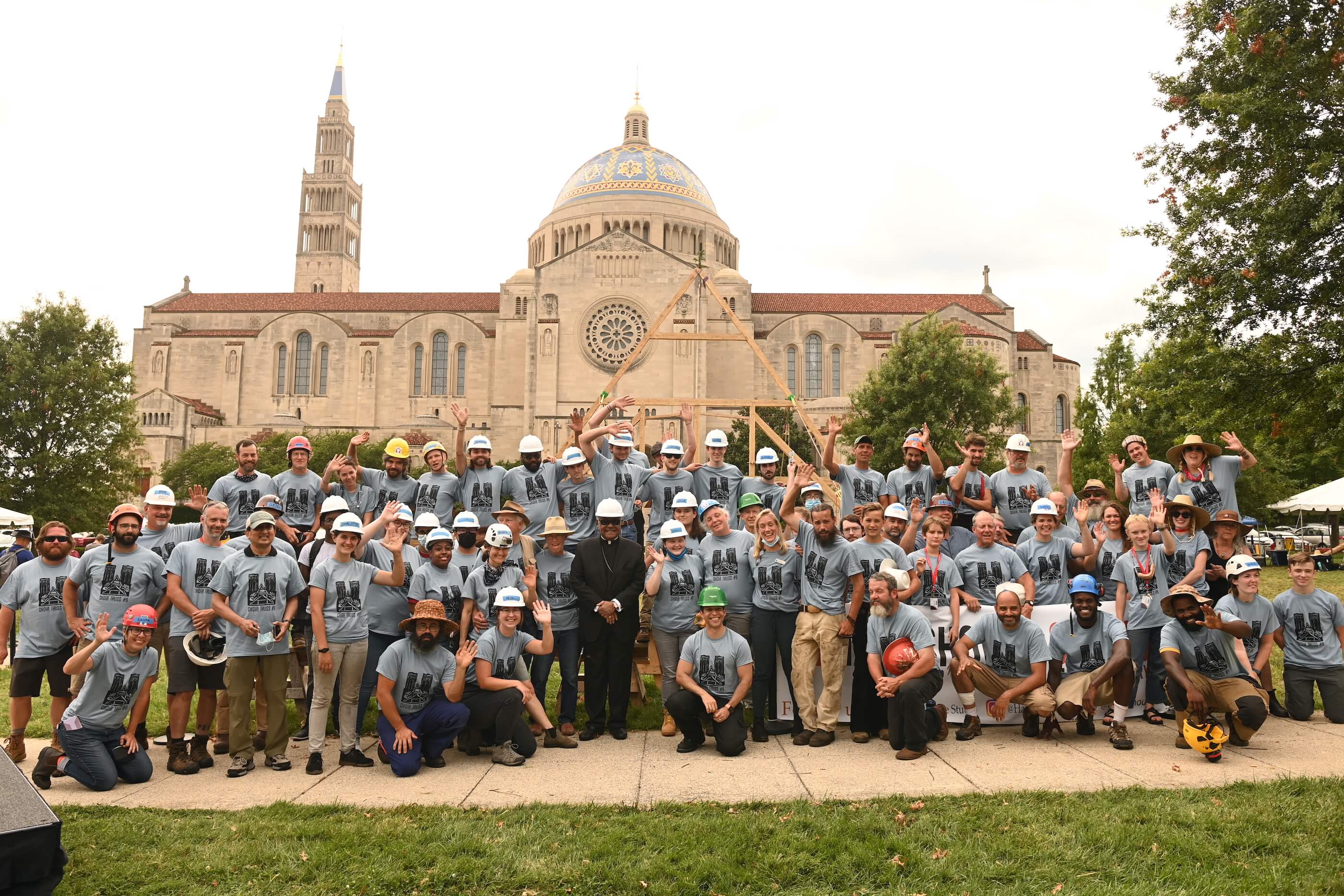 a team poses in front a wooden truss installed outdoors in front of a basilica
