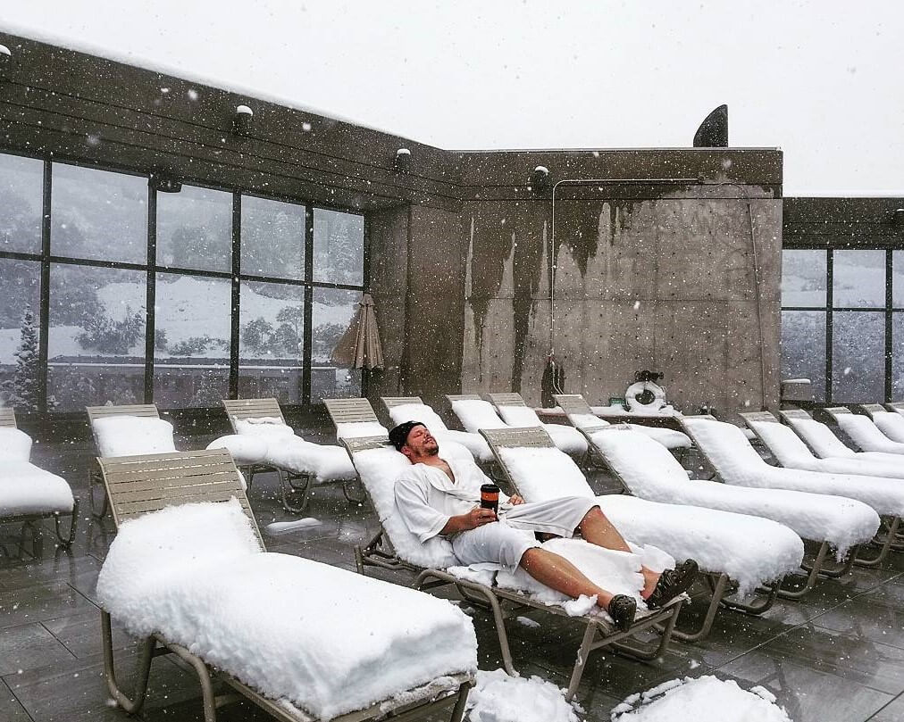 an outdoor terrace with lounge chairs covered in snow and a person laying down in a robe with a cup of coffee