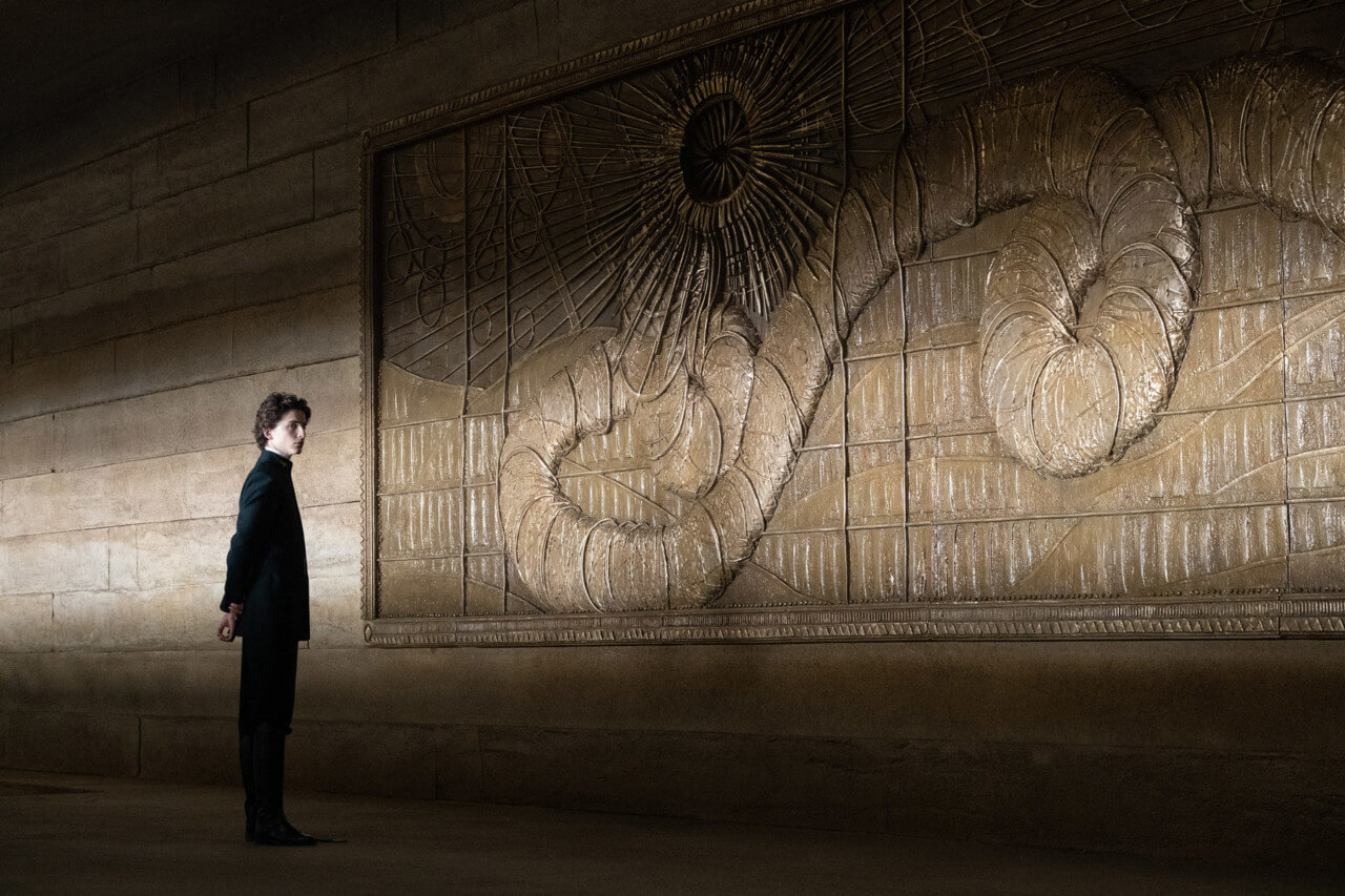A still from the 2021 dune film of a young man in front of a massive golden bas relief of a worm god