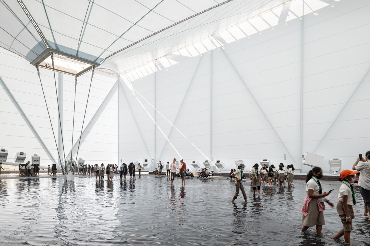 visitors wade across a pool of water in a hangar-like textile structure
