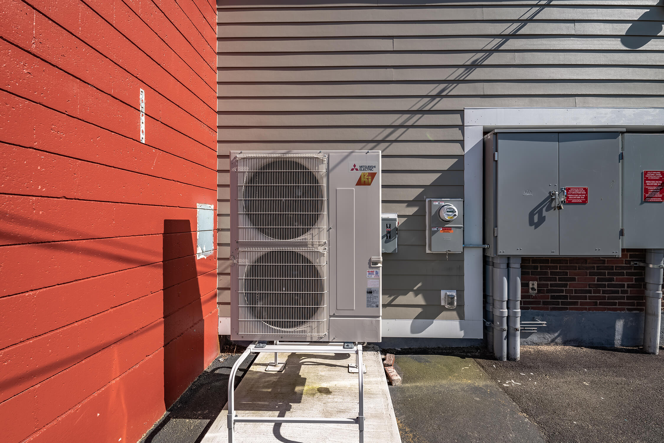 An AC unit in the corner of a building with red and gray shingles
