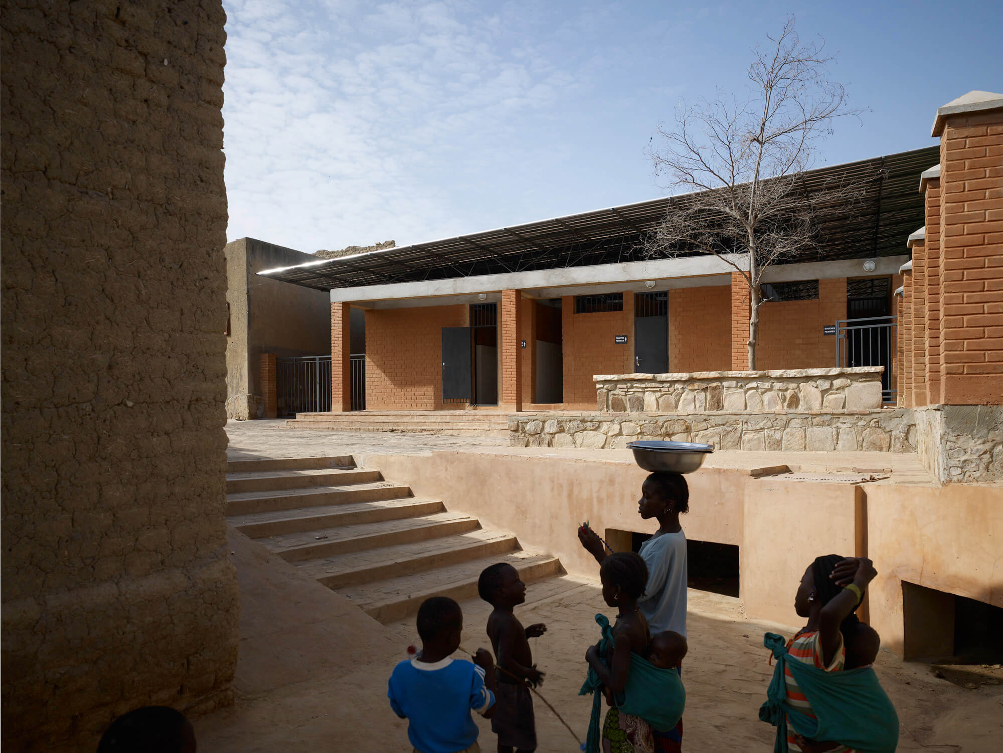 people gather in a shaded courtyard outside an earthen building