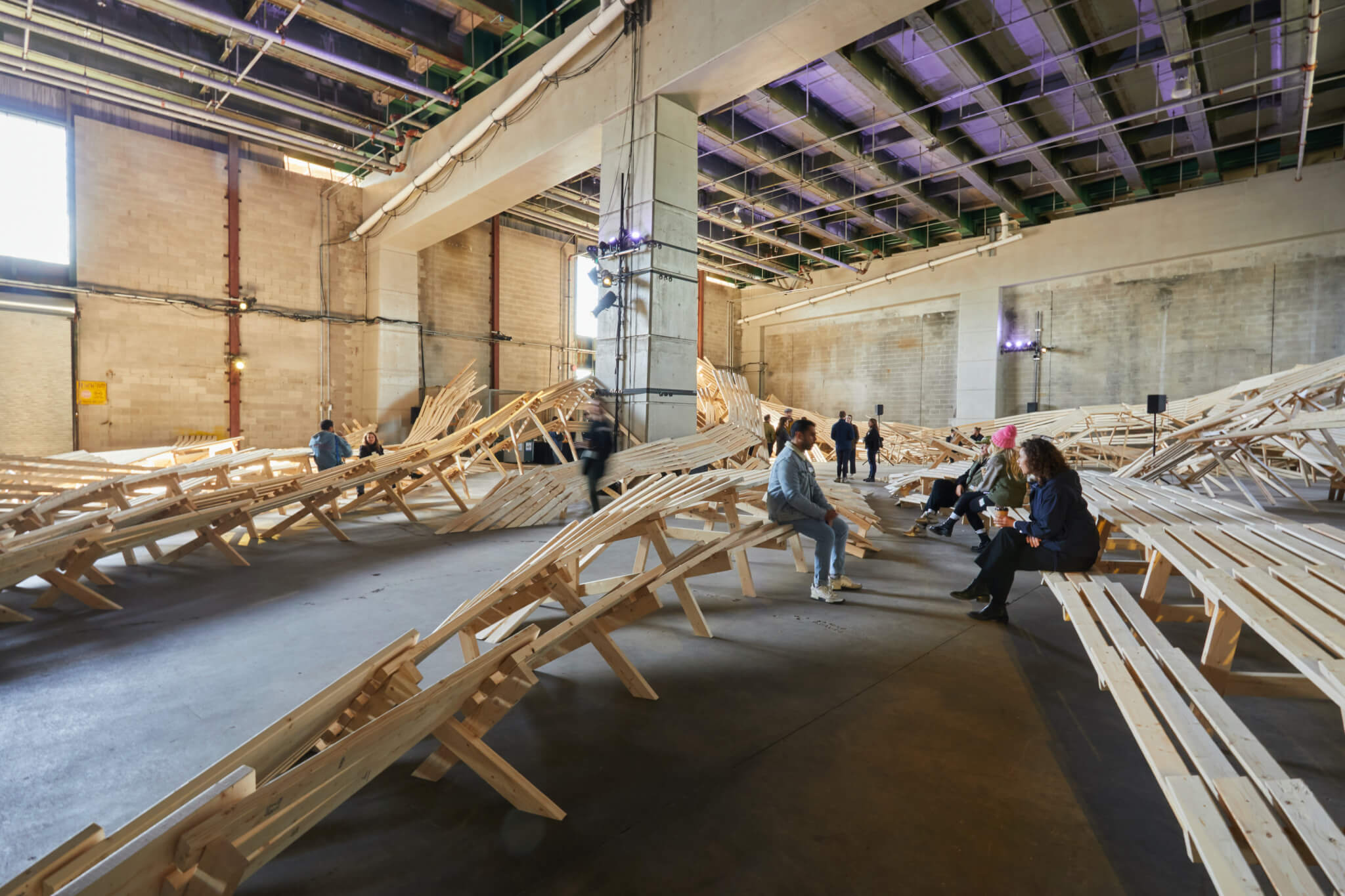 people sit on undulating wooden forms in a large open storage room