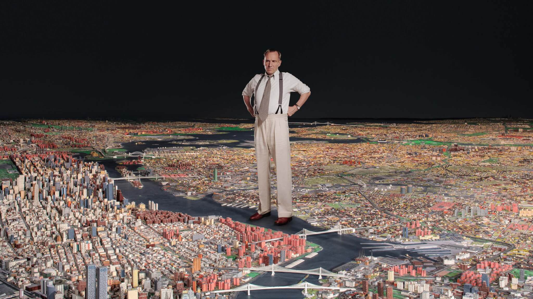 ralph fiennes as robert moses standing on model of new york city