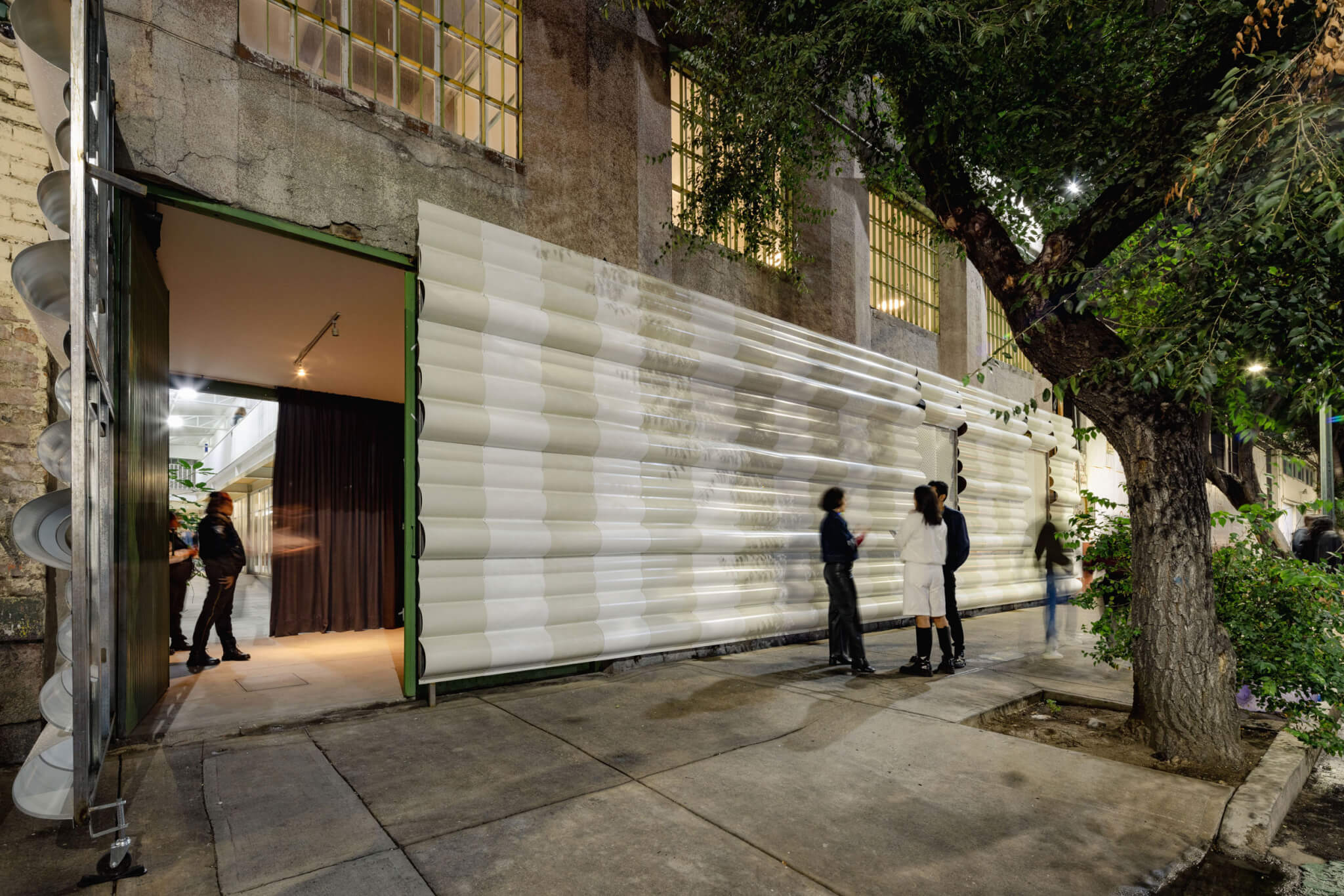 A white and beige striped facade underneath a warehouse building. The door to the studio is open, revealing the contrast between the outside and inside. People passing by converse by the tree in front of the building.