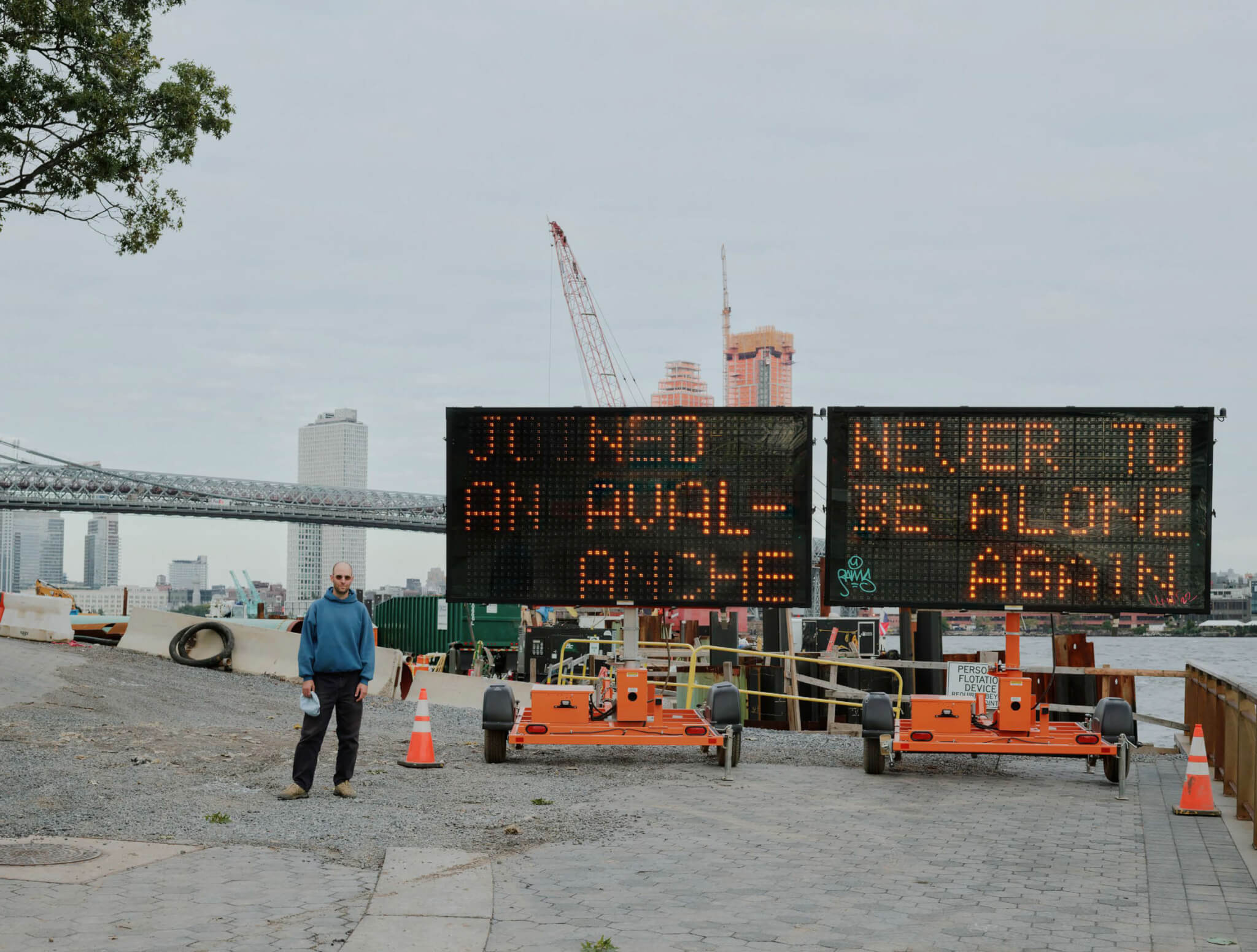 Man stands next to a construction sign that reads "Joined an avalanche, never to be alone again" overlooking the East River.
