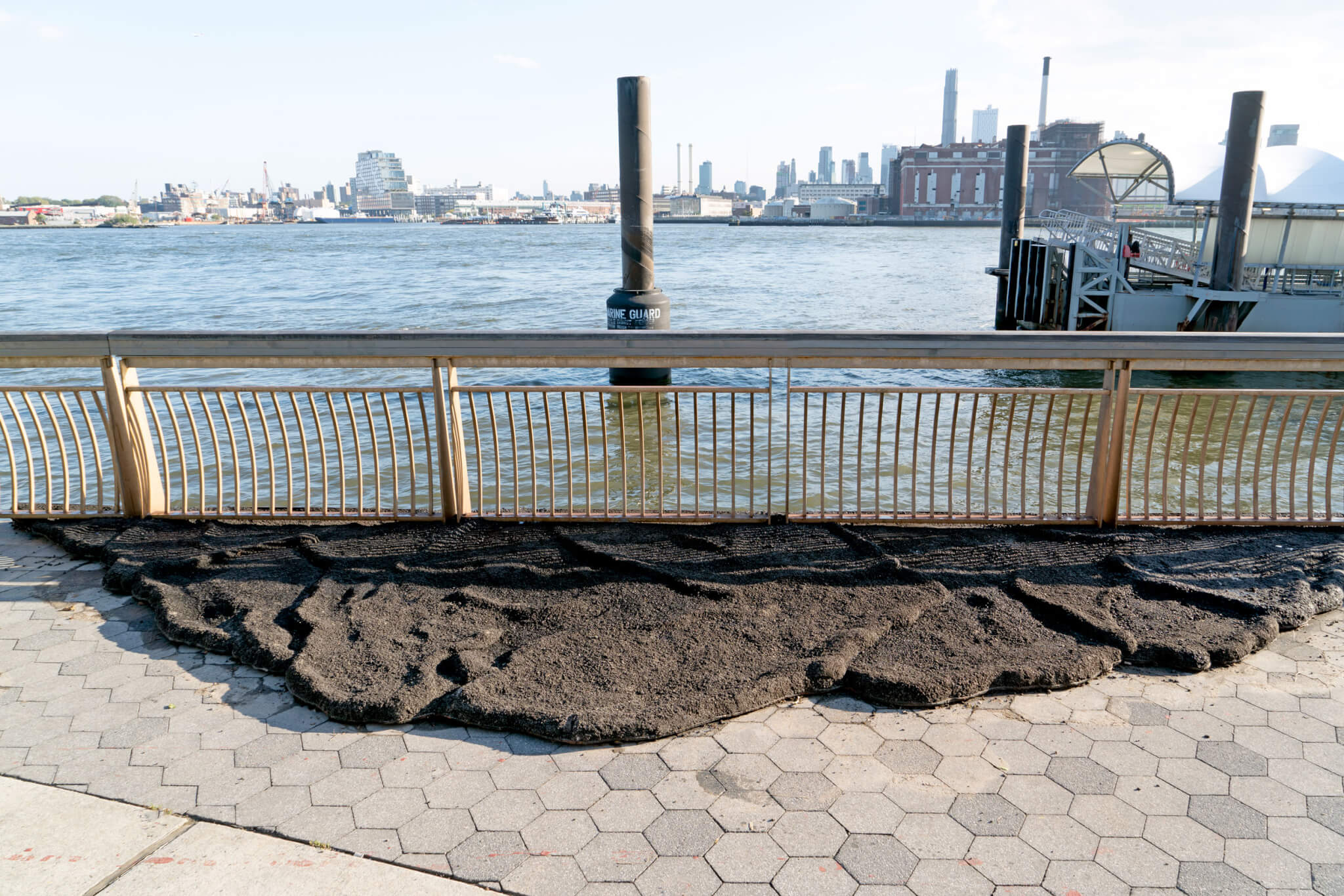 Photo of installation, black asphalt sculpture made to look like a tidal wave emerging from the wall barrier of the river.