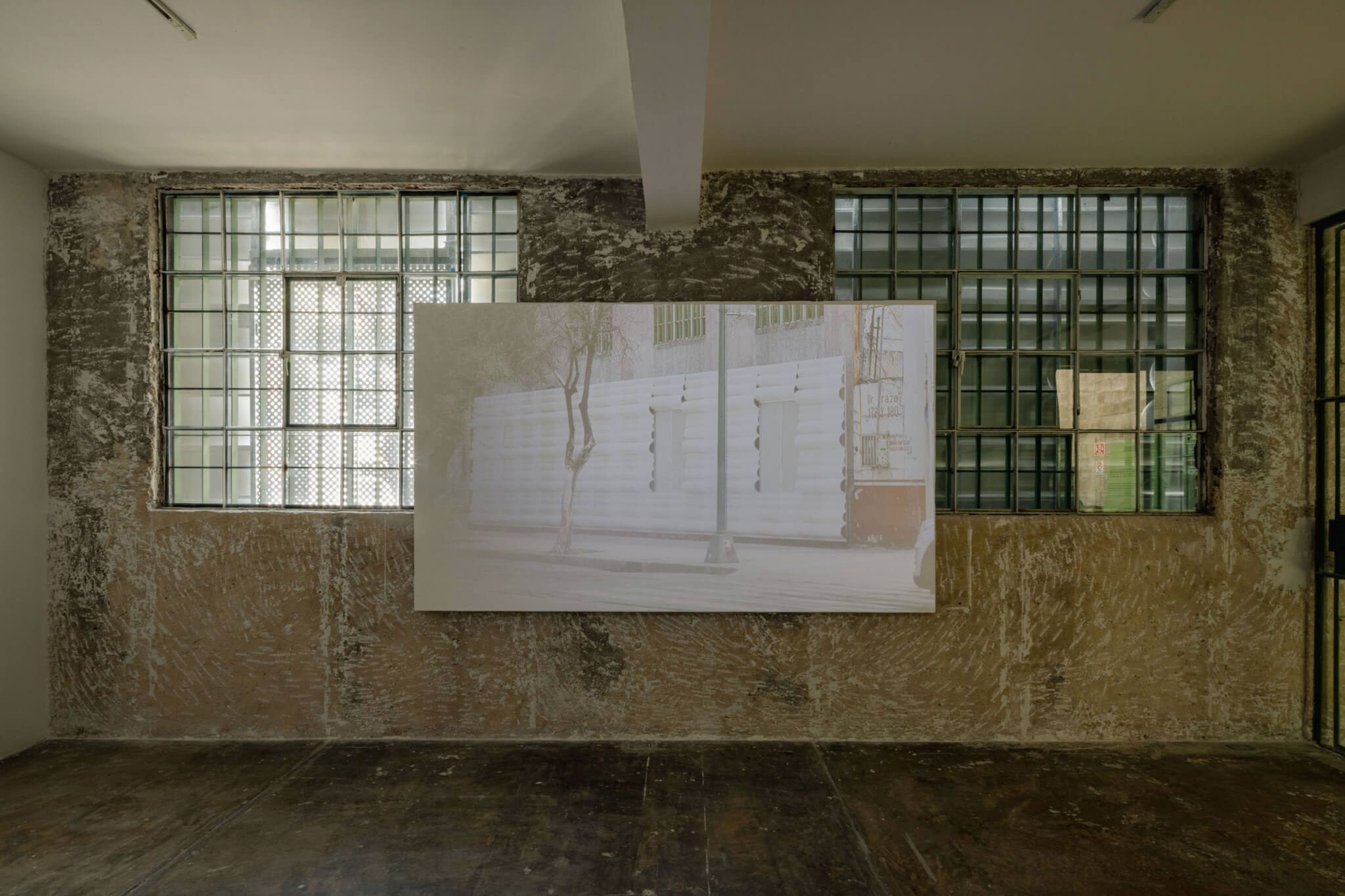 Room with two windows with the view obstructed by an object outside. A screen is in the middle of the room with an image of the outside's facade projected unto it.