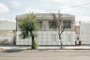 Image of LIGA 37’s gallery, an old warehouse surrounded by seemingly abandoned buildings. The gallery's facade is an installation, it covers the bottom half of the building with curved white and beige lines. Two trees lay in front of the building.