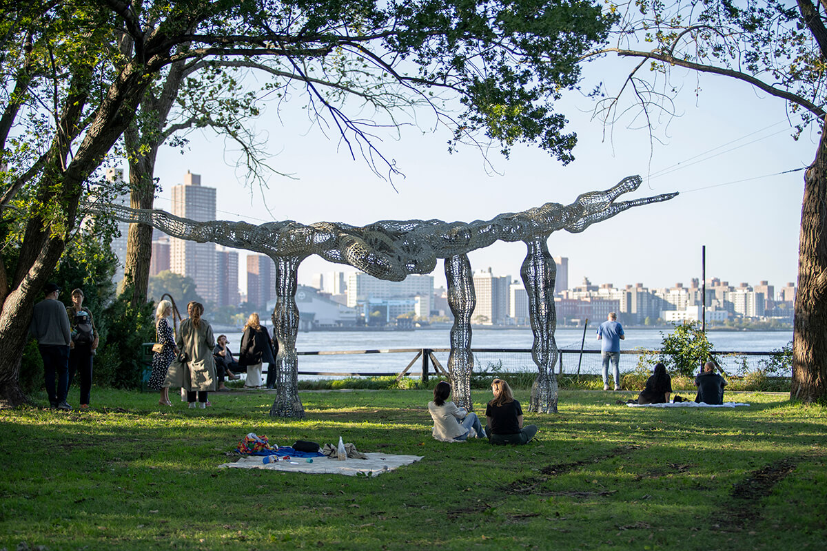 Image of an organic looking aluminium sculture in a park overlooking the river, with people sitting around the sculpture.