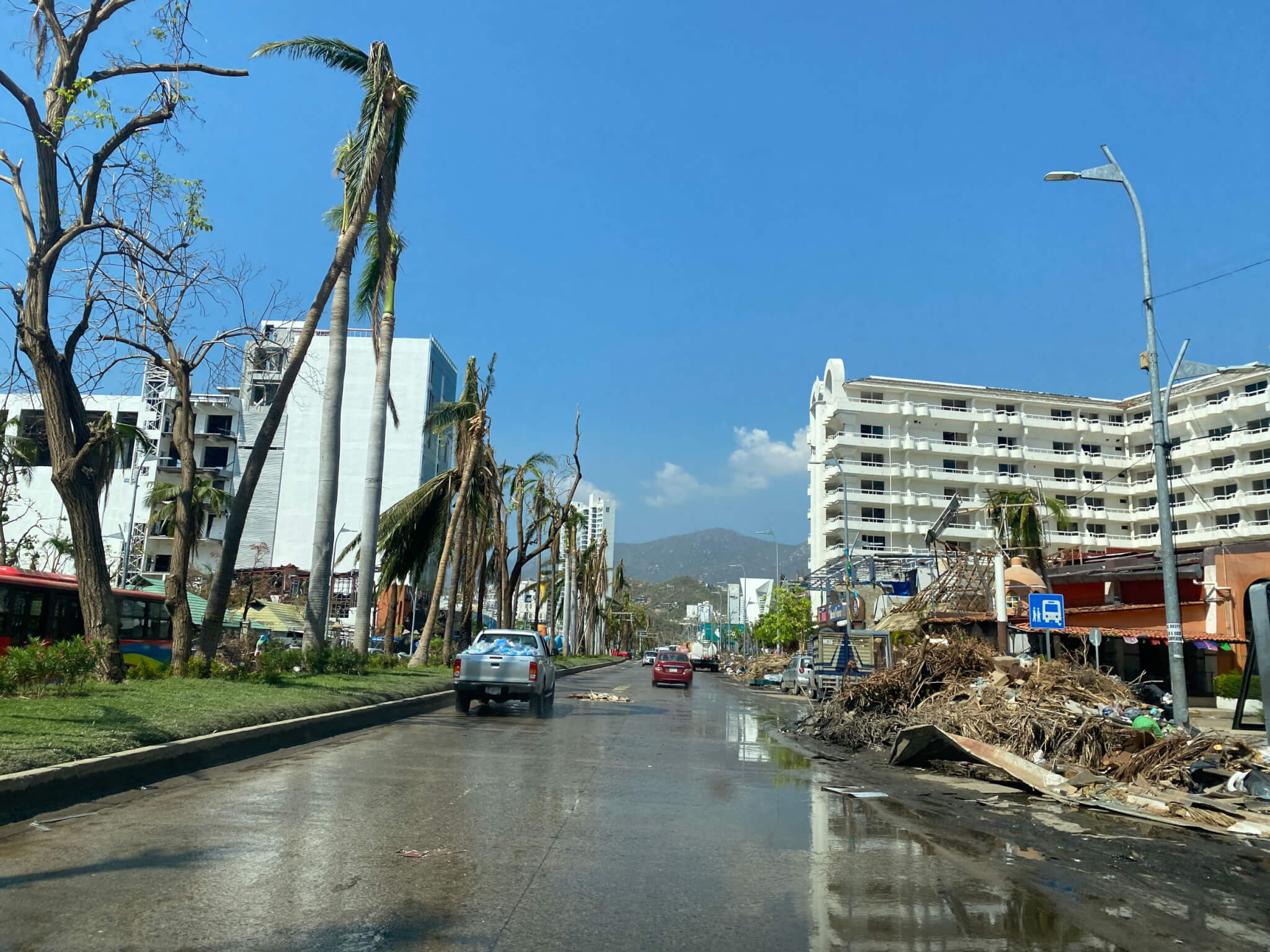 A street with damaged palm trees on the left and a damaged short structure to the right, with a standing white building on the background.